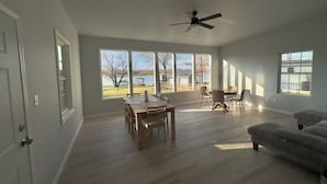 Amplia sala de estar con un gran ventanal con vista a un lago, comedor con mesa y sillas, y un sofá gris.