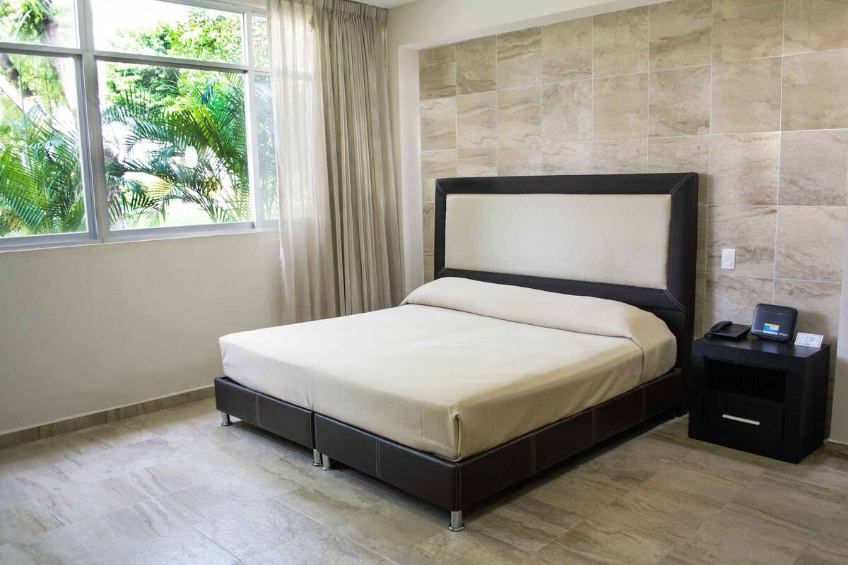 A modern hotel room with a large bed, a bedside table with a phone and a charging station, a window with curtains, and a tiled wall.