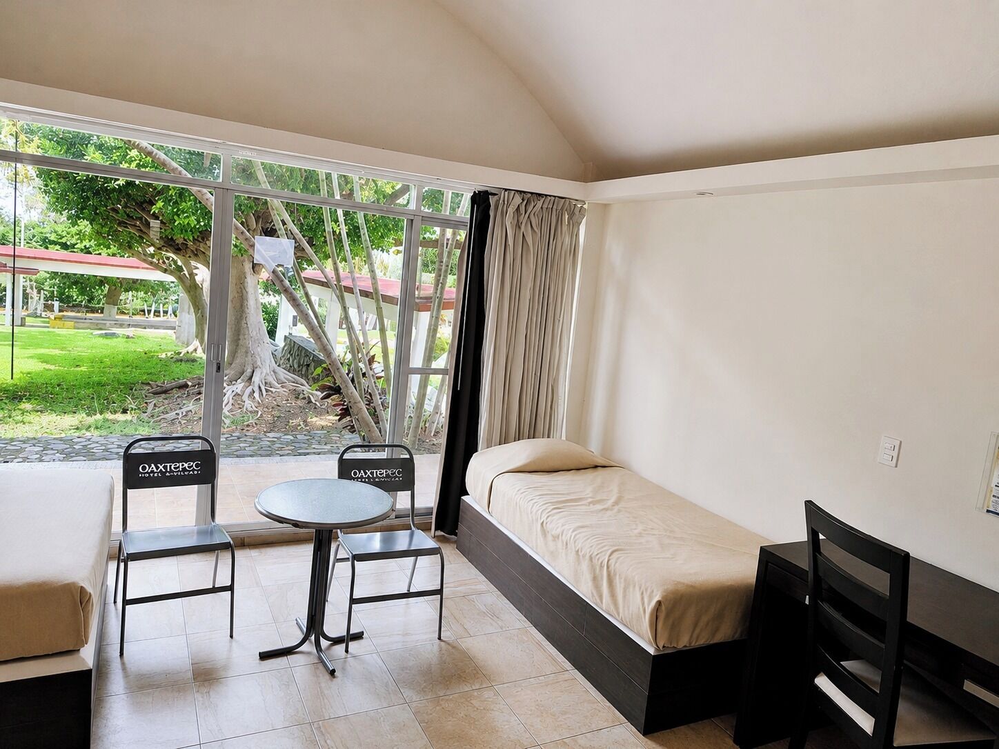 A small, single-room hotel room with a bed, a table, two chairs, and a window with curtains.