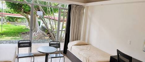 A small, single-room hotel room with a bed, a table, two chairs, and a window with curtains.