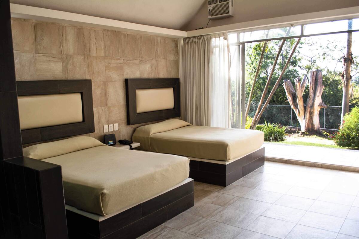A hotel room with two beds, a large window, and a view of greenery.
