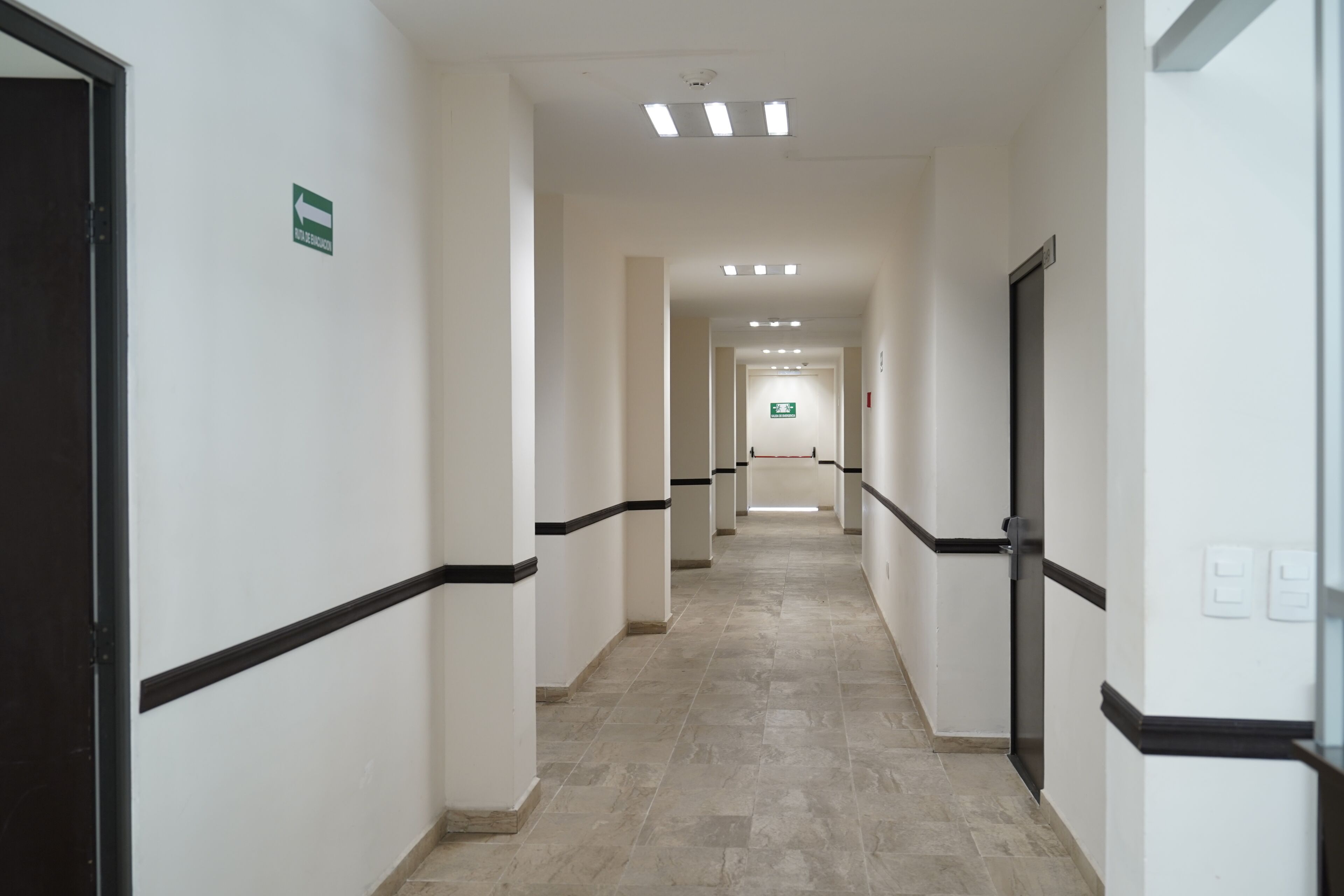Un pasillo con paredes blancas, detalles en negro y suelo de baldosas, que conduce a una señal de salida.