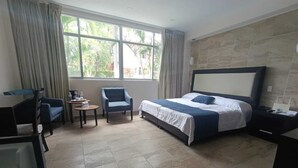 Quarto de hotel com cama, escrivaninha, cadeira, mesinha e janela com cortinas.
