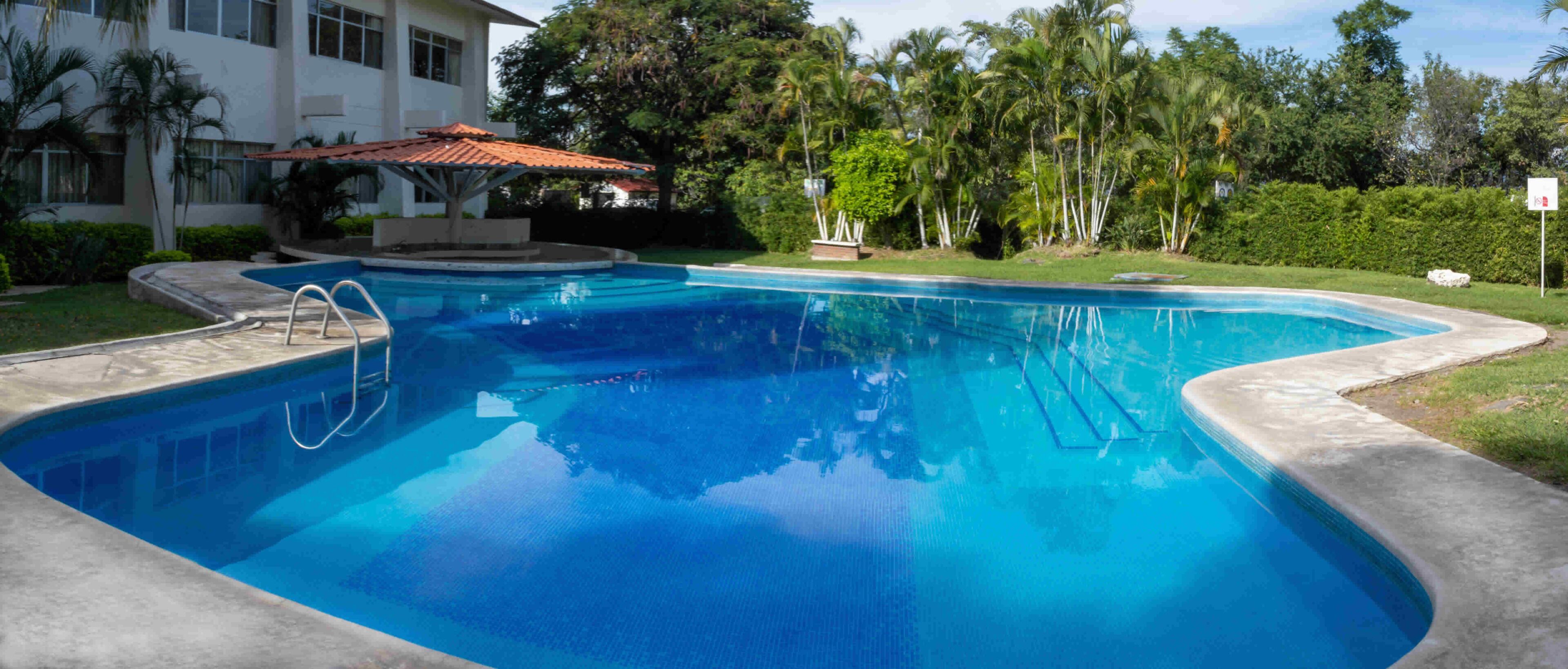 A swimming pool with a tiled deck, a covered seating area, and lush greenery surrounding the property.