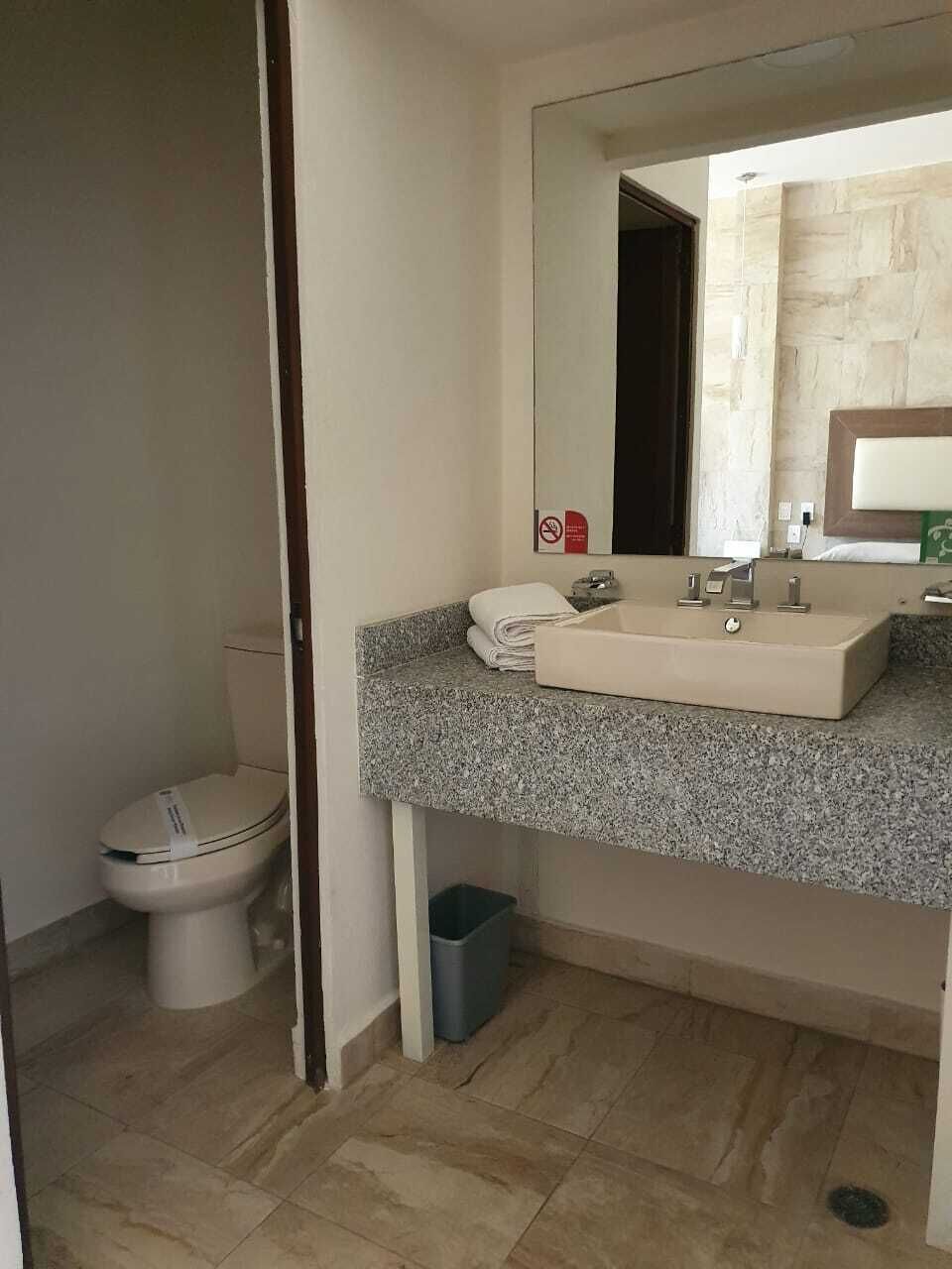 Un baño con inodoro blanco, lavamanos de encimera de granito, espejo y un cubo de basura.