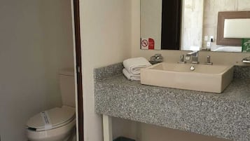 Un baño con inodoro blanco, lavamanos de encimera de granito, espejo y un cubo de basura.
