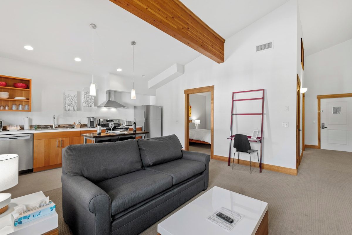 A modern living space with a grey sofa, a white coffee table, and a kitchen area with wooden cabinets and stainless steel appliances.