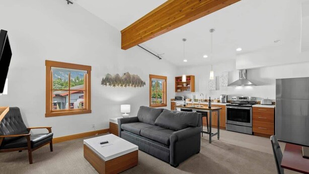 A modern living room with a grey sofa, a coffee table, and a TV. There is a kitchen area with a stove, oven, and cabinets. - Walk to Village-Fireplace-Festhalle Section (Leavenworth)
