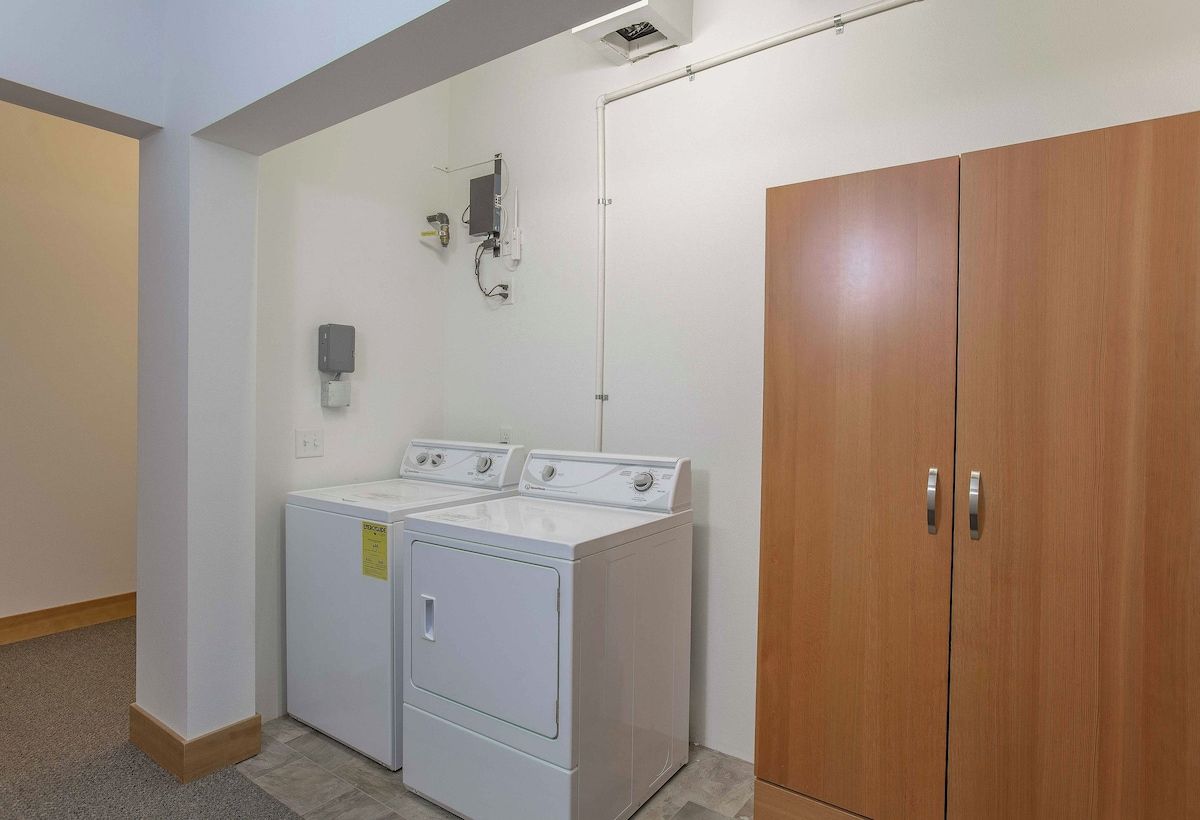 Een wasserette met twee witte wasmachines en drogers, een houten garderobe en een elektrische verdeeldoos aan de muur.
