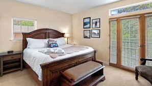 Quarto com cama grande, mesa de cabeceira, um banco, janela com persianas e porta com vista.