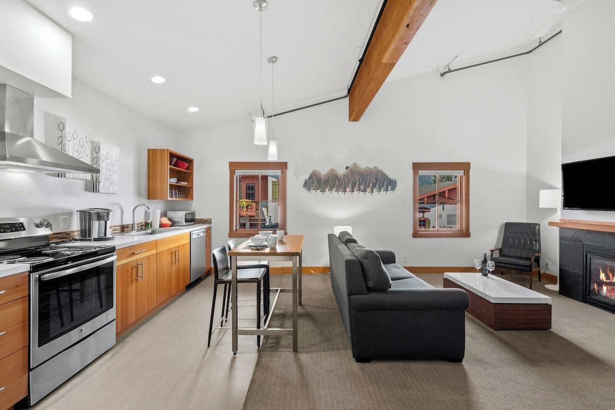 Een moderne keuken met roestvrijstalen apparatuur, een eetgedeelte met een tafel en stoelen, een woonkamer met een bank en een open haard, en kunstwerken aan de muren.