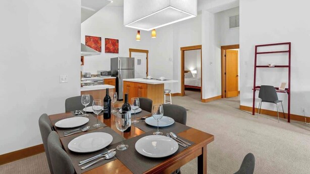 Ein zeitgemäßer Essbereich mit einem Holztisch, gedeckt für vier Personen, einer Küche im Hintergrund und einem roten Metallregal an der Wand.