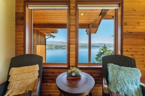 A room with two wicker chairs, a small round table, and a view of a lake through large windows.