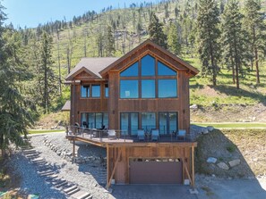 A two-story wooden cabin with a large balcony, surrounded by a forested landscape.