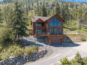A two-story wooden cabin with a large balcony, surrounded by a gravel driveway and a stone wall.