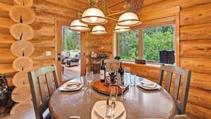 Uma área de jantar em uma cabana de madeira com uma mesa redonda posta para quatro pessoas, cercada por cadeiras de madeira. Um lustre pendurado no teto e uma janela oferecem vista para a floresta lá fora.