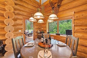 A wooden cabin dining area with a round table set for four, surrounded by wooden chairs. A chandelier hangs from the ceiling, and a window provides a view of the forest outside. - Private lodge with stunning mountain views (Leavenworth)