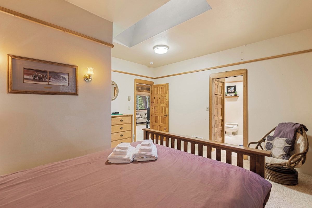 A bedroom with a bed, a wooden bench, a chair, a framed picture on the wall, and a door leading to a bathroom.