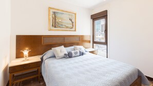 Ein ordentlich eingerichtetes Schlafzimmer mit einem großen Bett, einem Nachttisch mit Lampe, einem gerahmten Kunstwerk an der Wand und einem Fenster mit Aussicht.