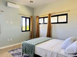 A bedroom with a bed, a window with curtains, an air conditioning unit, and a rug. - Coastal at Haven+ (St.kitts)
