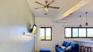 A modern living room with a blue sofa, a ceiling fan, and a large window. - Coastal at Haven+ (St.kitts)