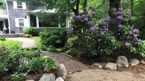 Un jardin avec un chemin en briques, de gros rochers et des arbustes aux fleurs violettes.