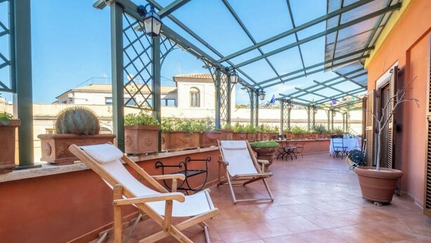 Een overdekt buitenzitgedeelte met lounge fauteuils en tafels, omringd door terracotta tegels en potplanten.