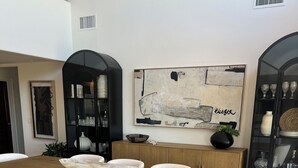 Uma zona de refeições moderna com uma mesa de madeira e cadeiras brancas, um vaso preto com folhagem e um quadro abstrato grande na parede.