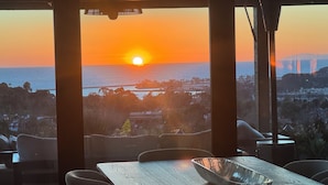 Uma área de refeições com vista para o pôr do sol sobre o mar, uma mesa de madeira e cadeiras modernas.