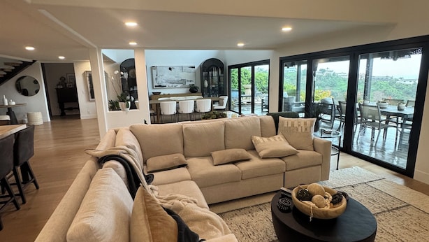 Uma sala de estar espaçosa com um amplo sofá modular bege, uma mesa de centro preta e vista para o exterior através de grandes portas de correr de vidro.