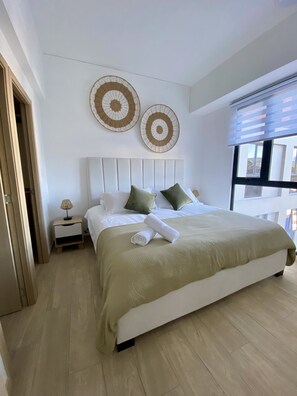 Un dormitorio moderno con una cama grande, una mesita de noche, dos obras de arte tejidas colgadas en la pared y una ventana con persianas.