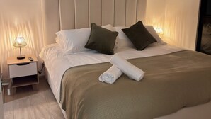 Una cama bien hecha con una colcha blanca y un plaid verde, una lámpara de noche y una mesita de noche de madera con un cajón.