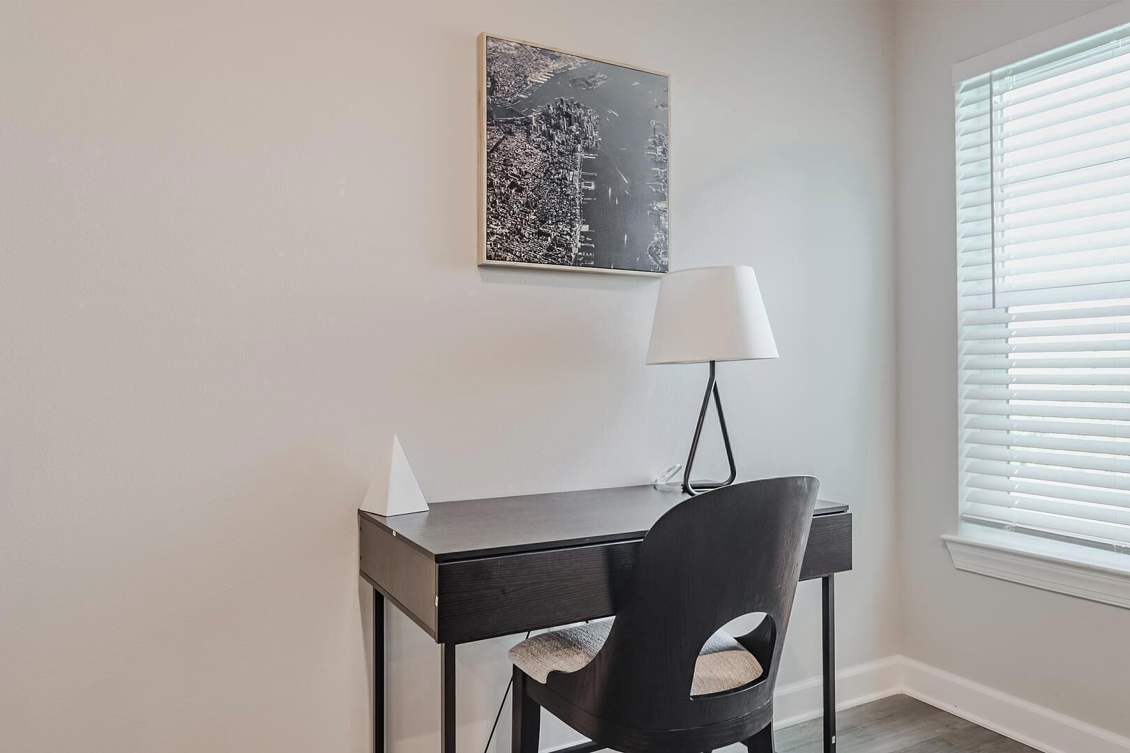 Ein kleiner Büroraum mit Schreibtisch, Stuhl, Lampe und einem gerahmten Stadtbild an der Wand.
