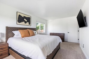 A modern bedroom with a large bed, a nightstand, a dresser, a television, and a window with a view of greenery. - Landing | Stunning 3BD, Clubhouse, Gym (Billings)