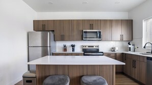 Eine moderne Küche mit Holzschränken, einem Edelstahlkühlschrank, einer Mikrowelle, einem Backofen und einem Geschirrspüler. An der Kücheninsel stehen zwei Barhocker.