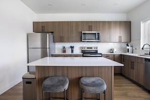 A modern kitchen with wooden cabinets, a stainless steel refrigerator, a microwave, an oven, and a dishwasher. There are two bar stools at the island. - Landing | Modern 3BD, Gym, Clubhouse (Billings)