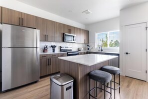 A modern kitchen with stainless steel appliances, wooden cabinets, a central island with bar stools, and a window providing a view. - Landing | Beautiful 3BD, Clubhouse, Gym (Billings)