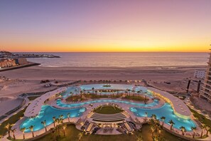 A resort with a large, winding pool surrounded by palm trees and lounge areas, overlooking a beach and ocean at sunset.