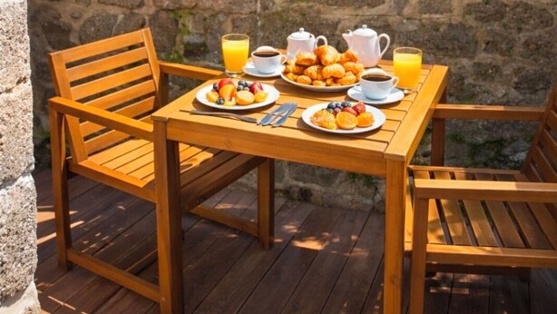 Une table en bois, dressée pour le petit-déjeuner, avec des fruits, des viennoiseries et des boissons, entourée de chaises sur une terrasse. Un mur de pierre se trouve en arrière-plan.