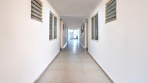 A long, narrow hallway with white walls and a tiled floor, flanked by windows with blinds. - Classic Royal Rooms (Bengaluru)
