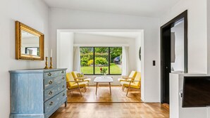 Ein Zimmer mit einem blauen Kommode, zwei gelben Stühlen, einem weißen Tisch und einem großen Fenster mit Blick auf Grünflächen und ein Auto davor.