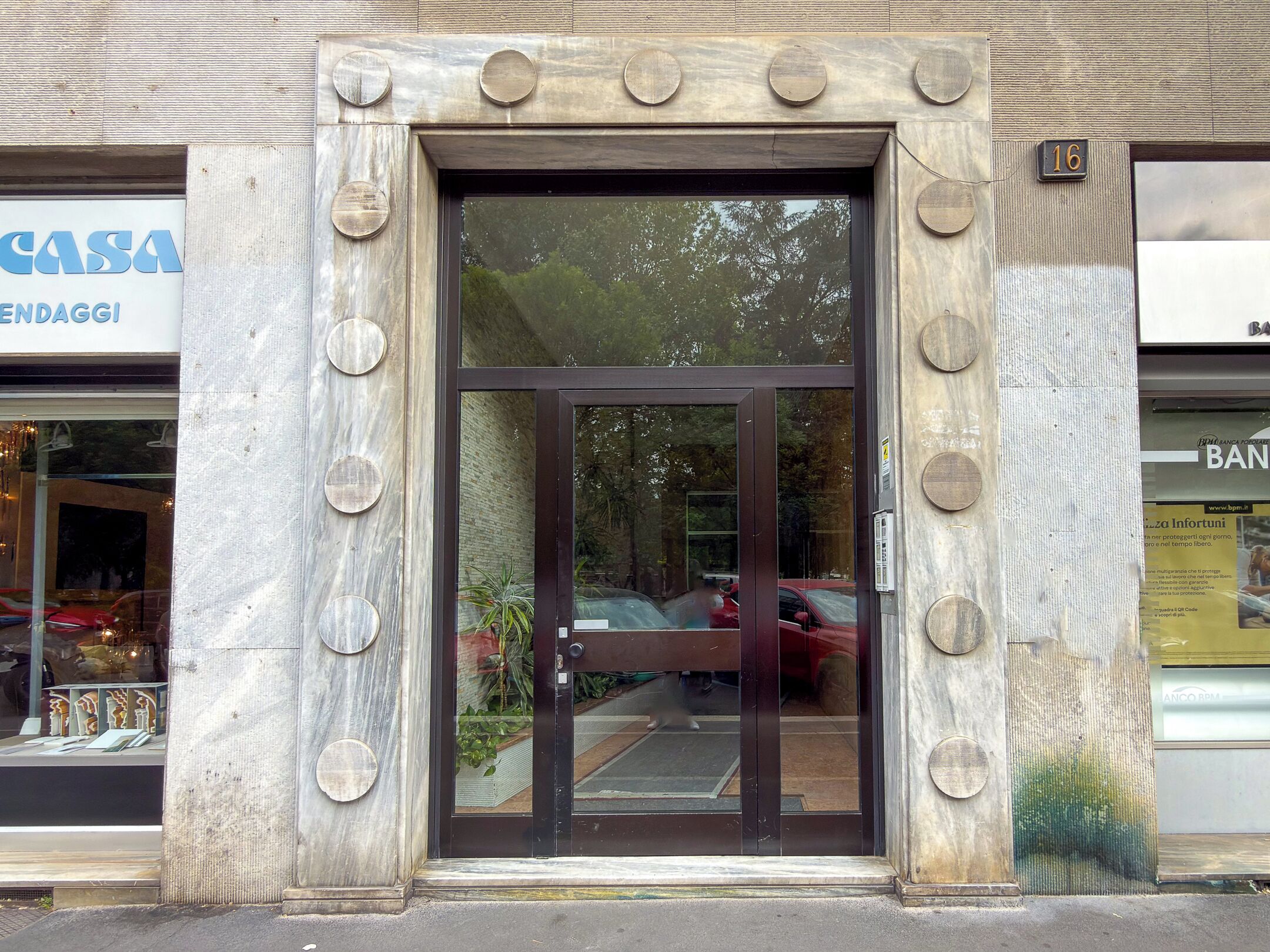 Une entrée d’immeuble avec une porte en verre, une enseigne indiquant « CASA EN DAGGI » et une pancarte « BAN » sur la droite.