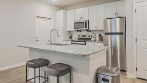 Una cocina moderna con gabinetes blancos, electrodomésticos de acero inoxidable, encimera de granito y dos taburetes de bar.