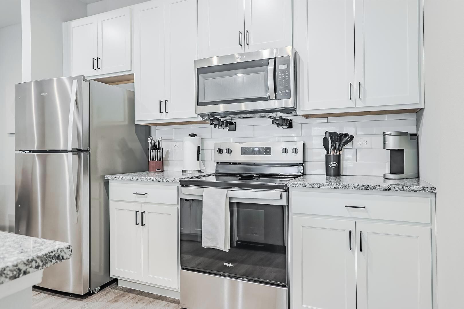 A modern kitchen with white cabinets, stainless steel appliances including a refrigerator, microwave, and oven, and a granite countertop.