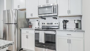 Una cocina moderna con gabinetes blancos, electrodomésticos de acero inoxidable, incluyendo un refrigerador, microondas y horno, y encimera de granito.