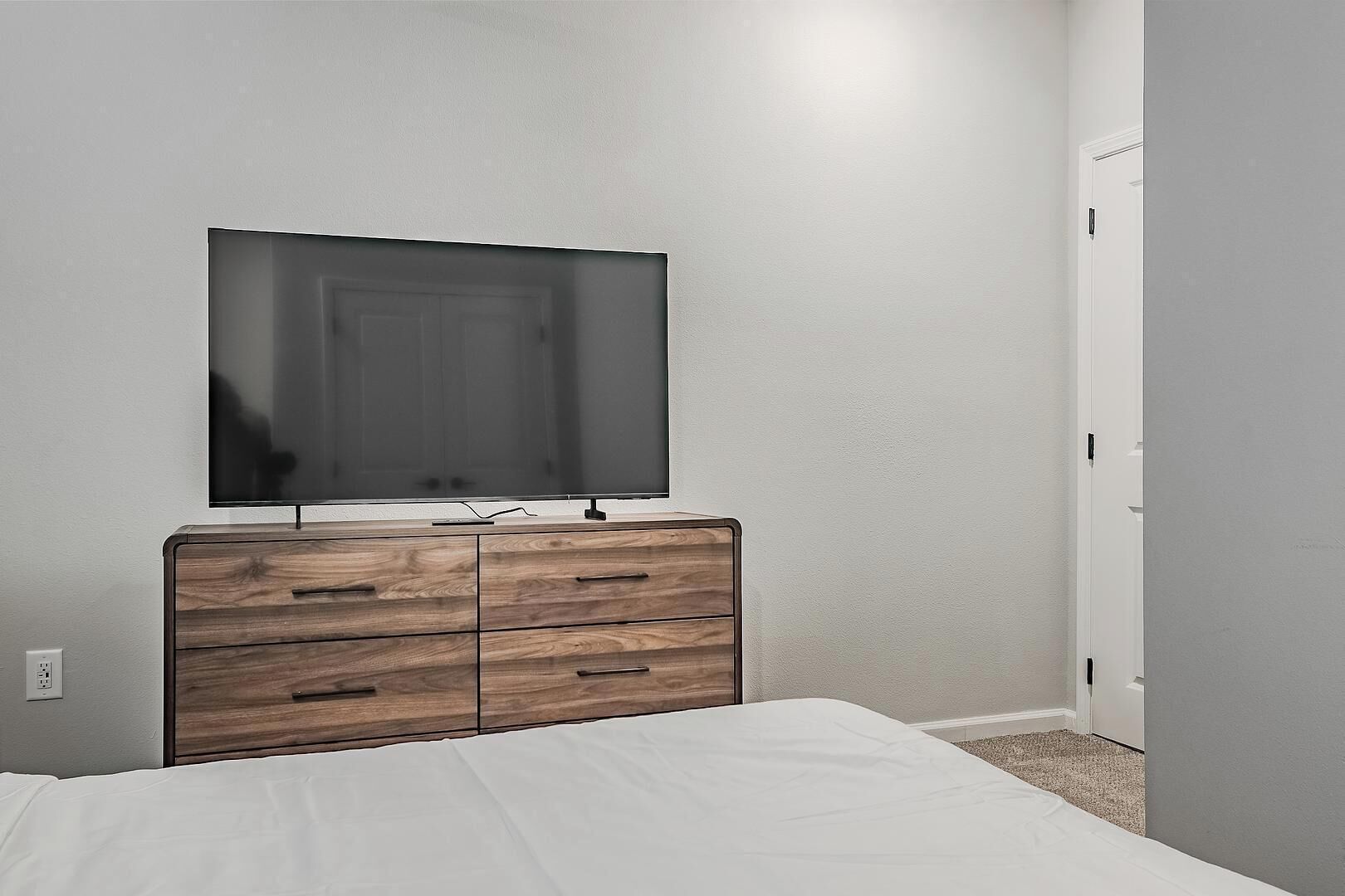 Quarto com cama, cómoda de madeira com gavetas, TV de ecrã plano fixada acima da cómoda e porta branca à direita.