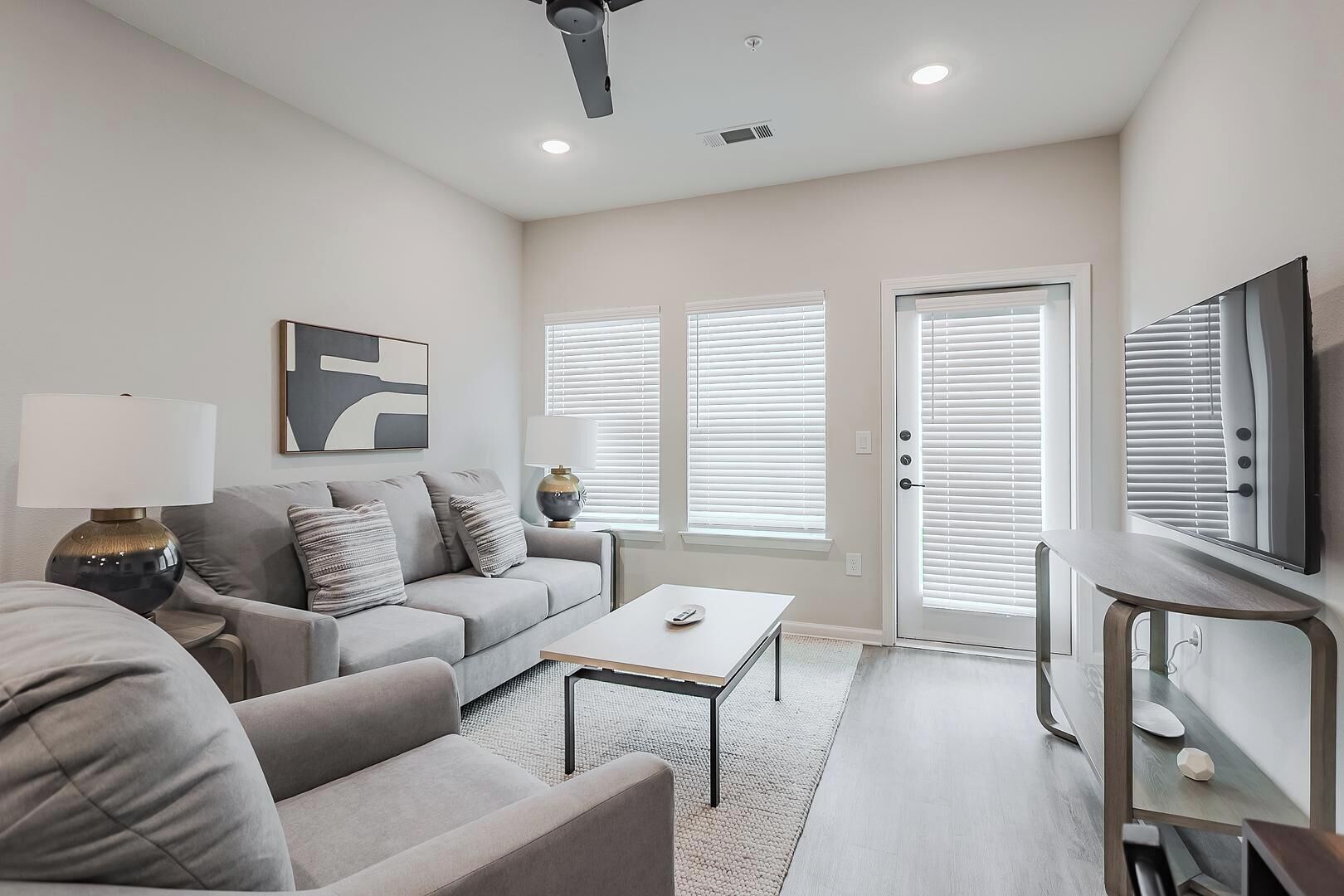 A modern living room with a grey sofa, a coffee table, a TV on a stand, and a door with blinds.