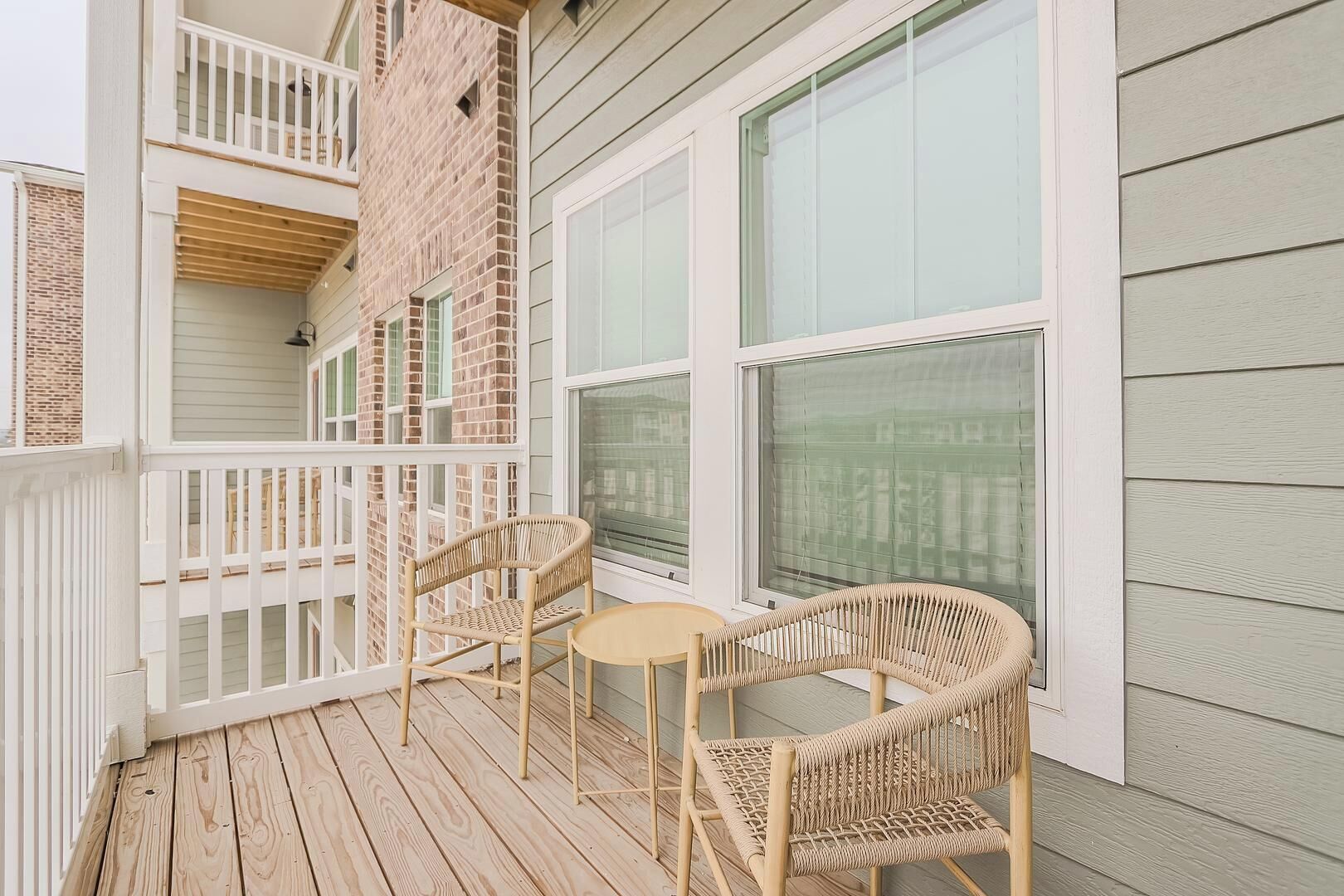 A wooden deck with two wicker chairs and a small table, overlooking a brick building with large windows.