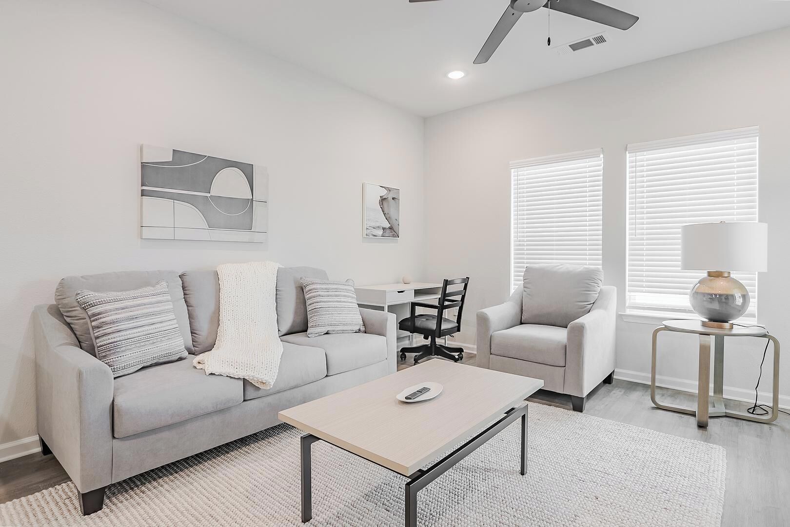 Un salon moderne avec un canapé gris, une table basse, une table d’appoint avec une lampe et un fauteuil. Il y a deux fenêtres avec des stores, un ventilateur de plafond et des œuvres d’art accrochées aux murs.