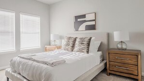 Un dormitorio con cama, dos mesitas de noche, una lámpara de pared y una ventana con persianas.